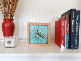 Square clock framed with cherry wood and patina copper face with pine cone design on a bookshelf.