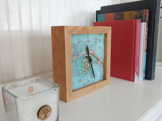 Square clock framed with cherry wood and patina copper face with pine cone design on a bookshelf.