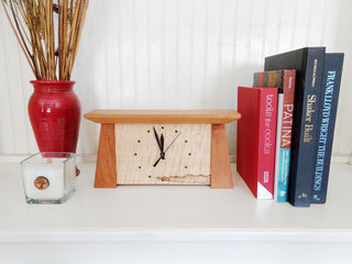 Tapered rectangular cherry wood framed maple wood clock on white shelf with books, vase, candle.