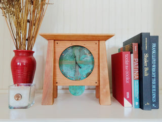 Cherry wood clock, 4 tapered legs, patina copper face & pendulum on white shelf with books & vase.
