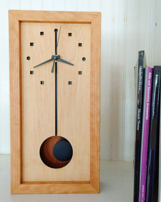 Vertical rectangular cherry wood clock with black pendulum on white shelf with books.