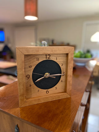 Leather & Cherry, handcrafted cherry wooden box clock featuring a numbered cherry face with cutout revealing black leather circle in the center. Pictured on wooden surface in home.
