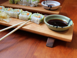 Close-up of sushi board and bowl with fresh sushi, soy sauce in white and blue bowl, and chopsticks.